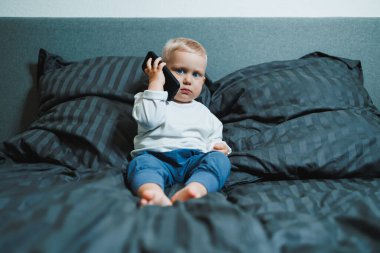 Akıllı telefonu olan 2 yaşında bir çocuk. Küçük çocuk yatakta oturuyor ve cep telefonuyla oynuyor. Annemi arıyorum. Elinde cep telefonu olan sevimli bir çocuk ekranda dikkatle bakıyor..