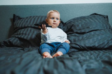 Küçük çocuk yatakta oturuyor, cep telefonuyla oynamaya çok odaklanmış. Tişört giyiyor ve telefonunda oyun oynuyor. Çocukların modern dijital hale getirilmesi. Cep telefonlu çocuk.