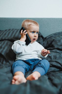 Küçük çocuk yatakta oturuyor, cep telefonuyla oynamaya çok odaklanmış. Tişört giyiyor ve telefonunda oyun oynuyor. Çocukların modern dijital hale getirilmesi. Cep telefonlu çocuk.