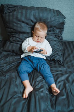 Küçük çocuk yatakta oturuyor, cep telefonuyla oynamaya çok odaklanmış. Tişört giyiyor ve telefonunda oyun oynuyor. Çocukların modern dijital hale getirilmesi. Cep telefonlu çocuk.