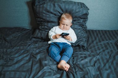 Küçük çocuk yatakta oturuyor, cep telefonuyla oynamaya çok odaklanmış. Tişört giyiyor ve telefonunda oyun oynuyor. Çocukların modern dijital hale getirilmesi. Cep telefonlu çocuk.
