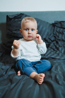 İki yaşında, mavi gözlü, uzun kollu beyaz gömlekli ve pantolonlu bir çocuk yatakta oturuyor. Yeni yürümeye başlamış çocuk yatak odasında, yatağın üstünde oturuyor..