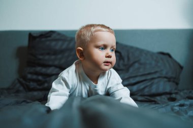 Çocuk yatakta. Beyaz bluzlu ve pantolonlu tatlı çocuk yatakta otururken gülüyor. 2 yaşında bir çocuk yatakta otururken oyun oynuyor. Erkekler için ev kıyafetleri.