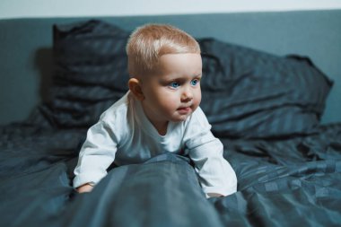 İki yaşında, mavi gözlü, uzun kollu beyaz gömlekli ve pantolonlu bir çocuk yatakta oturuyor. Yeni yürümeye başlamış çocuk yatak odasında, yatağın üstünde oturuyor..