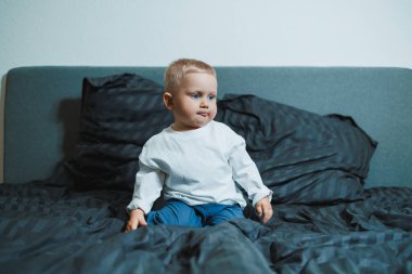 Çocuk yatakta. Beyaz bluzlu ve pantolonlu tatlı çocuk yatakta otururken gülüyor. 2 yaşında bir çocuk yatakta otururken oyun oynuyor. Erkekler için ev kıyafetleri.