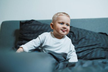 Beyaz gömlekli ve pantolonlu tatlı çocuk yatakta otururken gülüyor. 2 yaşında bir çocuk yatakta otururken oyun oynuyor. Erkekler için ev kıyafetleri.