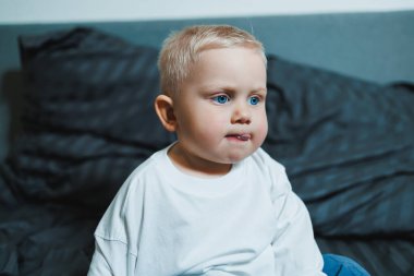 Çocuk yatakta. Beyaz bluzlu ve pantolonlu tatlı çocuk yatakta otururken gülüyor. 2 yaşında bir çocuk yatakta otururken oyun oynuyor. Erkekler için ev kıyafetleri.