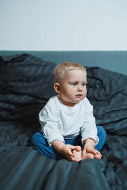 Çocuk yatakta. Beyaz bluzlu ve pantolonlu tatlı çocuk yatakta otururken gülüyor. 2 yaşında bir çocuk yatakta otururken oyun oynuyor. Erkekler için ev kıyafetleri.