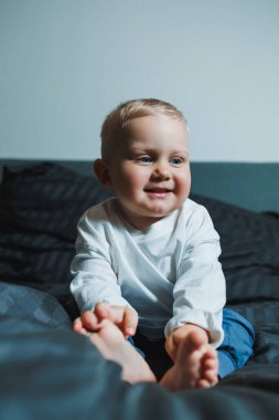 Beyaz gömlekli ve pantolonlu tatlı çocuk yatakta otururken gülüyor. 2 yaşında bir çocuk yatakta otururken oyun oynuyor. Erkekler için ev kıyafetleri.