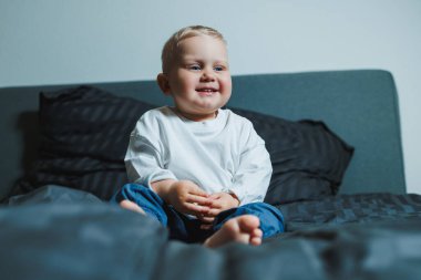 Beyaz gömlekli ve pantolonlu tatlı çocuk yatakta otururken gülüyor. 2 yaşında bir çocuk yatakta otururken oyun oynuyor. Erkekler için ev kıyafetleri.