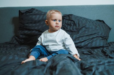 İki yaşında, mavi gözlü, uzun kollu beyaz gömlekli ve pantolonlu bir çocuk yatakta oturuyor. Yeni yürümeye başlamış çocuk yatak odasında, yatağın üstünde oturuyor..