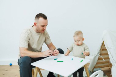 Babam 1.6 yaşındaki oğluna plastikten heykel yapmayı öğretiyor. Küçük çocuk babasıyla birlikte çocuk masasında oturur ve eğlenceli oyunlar oynar. Çocuk plastikten figürler yapıyor.