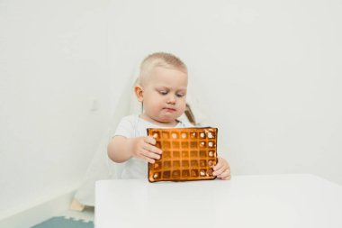 Merak ve merakla dolu küçük bir çocuk, mükemmel pişirilmiş waffle 'ı dikkatle inceler, basit, parlak ve neşeli bir ortamda keşfetme sevincini gösterir.