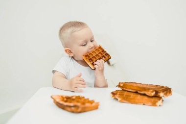 Çok tatlı ve neşeli bir çocuk, daha lezzetli waffle parçalarıyla çevrili beyaz bir masada otururken lezzetli bir waffle 'ı neşeyle ısırıyor.