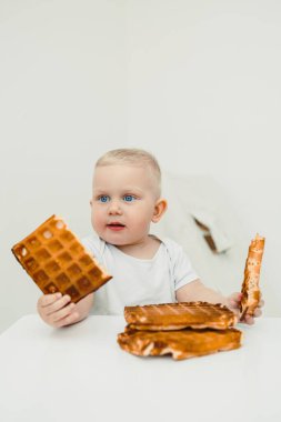 Parlak beyaz bir masada neşeli bir şekilde waffle parçaları tutan sevimli bir bebek. Güzel aile anları ve çocukluk anılarını yakalamak için mükemmel.