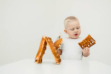 Kısa, açık renk saçlı küçük şirin bir çocuk iki farklı waffle şeklini inceliyor. Parlak, minimalist bir ortamda merak ve saf neşe hissi sergiliyor.