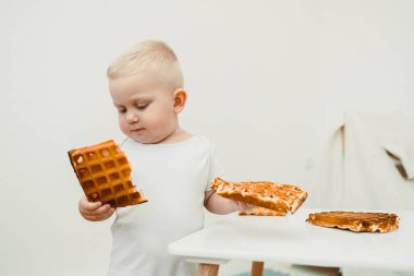 Çekici ve meraklı genç bir çocuk, basit, havadar bir mutfak ortamında keşif ve keşfin saf neşesini mükemmel bir şekilde somutlaştıran iki çıtır waffle 'ı heyecanla inceliyor.