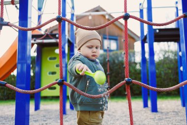 Güzel bir çocuk dışarıda oyun alanında eğleniyor, tırmanma ipleri ve renkli yapılarla etkileşime geçiyor, güvenli bir ortamda neşe ve keşif sergiliyor..