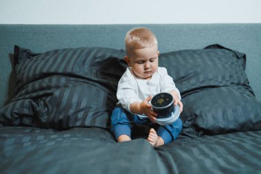 Kapalı bir yerde, neşeli bir çocuk yumuşak bir yatakta oturuyor, sürprizlerle dolu renkli bir oyuncak konteynırını keşfediyor, sıcaklık ve rahatlık için yumuşak yastıklarla çevrili.