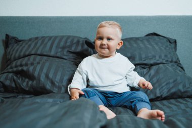 Neşeli bir çocuk rahat bir yatakta, koyu renkli yastıklarla oturur ve neşe saçar. Bu sahne aile içi ya da çocukluk dönemi için mükemmel.