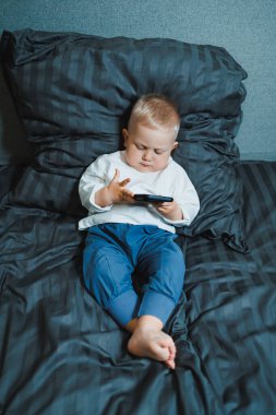 Genç bir çocuk rahat bir şekilde rahat bir yatakta, modern ebeveynlik eğilimlerini ve yeni yürümeye başlayan çocuklar için teknolojinin kullanımını vurgulayan akıllı bir telefona dalmış bir şekilde oturuyor.