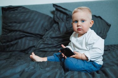 Sevimli bir bebek, rahat bir yatakta oturup, yumuşak yastıklarla çevrili akıllı bir telefonu inceliyor. Masum bir çocukluk keşfi için büyüleyici bir an.