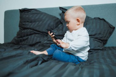 Meraklı bir çocuk, akıllı telefona odaklanmış bir şekilde yatakta oturuyor. Pofuduk yastıklarla çevrili çocuk, teknolojinin ve erken çocukluk keşiflerinin karışımını sergiliyor..