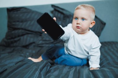 Sıcak bir atmosferde, meraklı bir çocuk yumuşak bir yatakta, elinde parlak bir akıllı telefon tutarak dijital çağda modern ebeveynlik ve çocukluk keşiflerini yakalıyor.