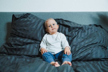 Beyaz gömlekli ve mavi pantolonlu çekici bir bebek yumuşak yastıklı rahat bir yatakta oturur, parlak, huzurlu ve neşeli bir ortamda masumiyet ve merak yayar.