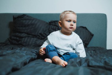 Kısa sarı saçlı şirin küçük bir çocuk rahat bir yatakta oturuyor, düşünceli bir şekilde etrafına bakıyor, şık ve modern yatak odasında koyu renkli yastıklarla çevrili.