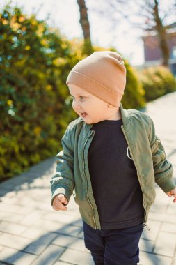 Neşeli bir çocuk güneşli bir günün tadını çıkarıyor. Havayı güzelleştiren canlı yeşilliklerle çevrili şık bir bombardıman ceketi ve rahat bir şapka giyiyor.