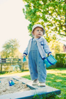 Renkli tulumlar içinde neşeli bir çocuk ve neşeli bir şapka oyuncaklarla dolu canlı bir kum havuzunda enerjik bir şekilde oynuyor, yemyeşil ağaçlar ve parlak mavi bir gökyüzü ile çevrili.