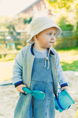 Rahat bir kıyafet giymiş neşeli bir çocuk güneşli bir günde etrafı renkli oyuncaklarla ve doğal güzelliklerle dolu canlı bir bahçeyle dolu bir kum havuzunu mutlulukla keşfeder.