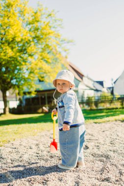 Güneşli güzel bir günde, neşeli bir genç çocuk verimli yeşillik, ağaçlar ve oyun malzemeleriyle çevrili parlak renkli bir kürekle kumlu bir bölgeyi mutlulukla keşfeder.