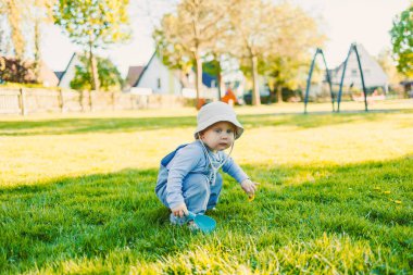Şirin şapkalı neşeli bir çocuk neşeli yeşil çimenlerin üzerinde çömeliyor, neşeli bir şekilde güneşli bir parkta oynuyor renkli salıncaklarla neşeli atmosferi güçlendiriyor.
