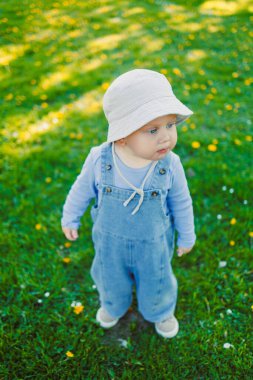 Kot pantolon giymiş ve güneş şapkası takmış şirin bir çocuk açık havada güneşli bir ortamda çocukluk sevincini somutlaştıran karahindiba desenli yeşil çimenlerin üzerinde duruyor..