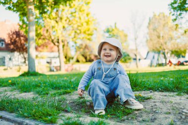 Parkta çimenlerde oturan, güneş şapkası takan ve dışarıda eğlenen mutlu bir çocuk. Çocukluk, eğlence ve yaz eğlencesi için mükemmel bir tema.