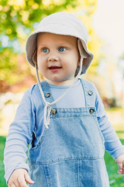Şirin şapkalı ve şık tulumlu çekici bir çocuk, neşeli bir parkta güneşli bir günün tadını çıkarıyor. Aile toplantıları ve hoş çocuklar için mükemmel bir fotoğraf.