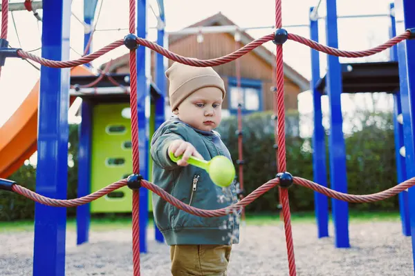 Güzel bir çocuk dışarıda oyun alanında eğleniyor, tırmanma ipleri ve renkli yapılarla etkileşime geçiyor, güvenli bir ortamda neşe ve keşif sergiliyor..