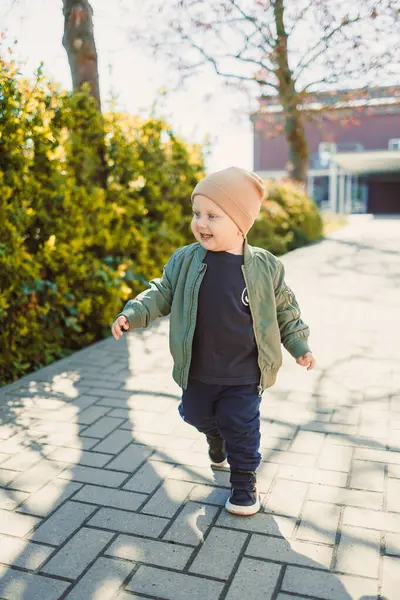 Parlak yeşil bir bombardıman ceketi ve rahat bir bere giyen neşeli bir çocuk, güneşli bir yolda yürüyor, gür çalılarla çevrili, çocukluk sevinci ve özgürlüğünü somutlaştıran
