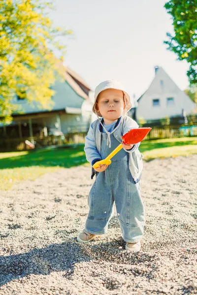 Renkli tulumlar içinde neşeli bir çocuk ve şirin bir şapka, kum havuzunda parlak kırmızı bir kürekle neşe içinde, çocukluk keşiflerinin neşesini somutlaştırıyor.