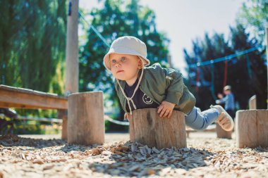 Güneşli bir günde, yeşil ceketli ve güneş şapkalı meraklı bir çocuk neşeli bir oyun parkında ahşap kütükleri keşfederek çocukluk ve aile eğlencesinin özünü somutlaştırıyor.