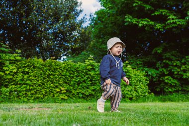 Neşeli bir çocuk, gülümsüyor, neşeli bir parkta neşeli bir şekilde yürüyor, şık bir kova şapkayla rahat bir kıyafet giyiyor, bu güzel günde doğanın güzelliğini kucaklıyor.