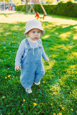 Mavi tulumlu sevimli bir bebek ve beyaz bir güneş şapkası renkli çiçeklerle dolu canlı yeşil bir parkta mutlu bir şekilde geziniyor, neşeli bir açık hava anını yakalıyor.