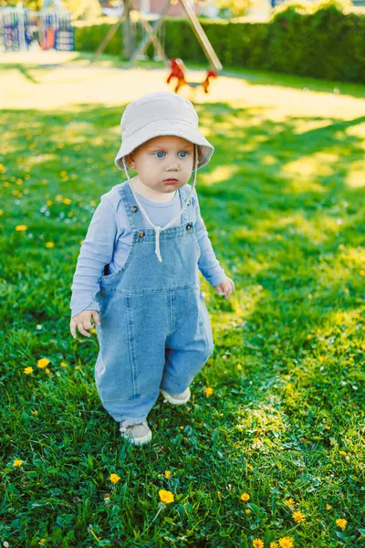 Mavi tulumlu sevimli bir bebek ve beyaz bir güneş şapkası renkli çiçeklerle dolu canlı yeşil bir parkta mutlu bir şekilde geziniyor, neşeli bir açık hava anını yakalıyor.