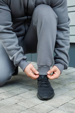 Bir insan gri spor ayakkabılarını giyerken kaldırımda diz çöker. Moda kıyafetlerini giyer. Heyecan verici bir aktivite ve egzersiz gününe hevesle hazırlanır.