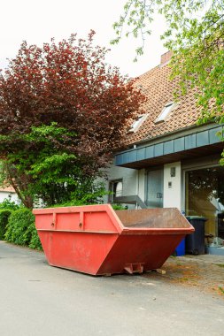 Parlak kırmızı bir çöp bidonu yeşil çalılar ve modern bir evle çevrili sessiz bir caddede duruyor, devam eden şehir inşaatını yansıtıyor ve bölgeye neşe katıyor.
