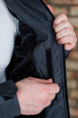 Bir kişi ustalıkla ve zarif bir şekilde iyi tasarlanmış bir ceket cebine uzanır, şık tasarım ve giysinin pratik özelliklerini canlı bir arka plana karşı sergiler.