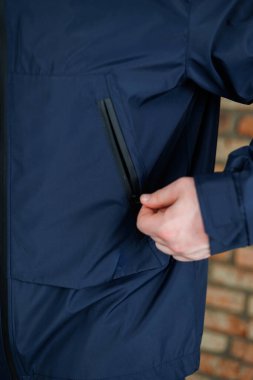 Detaylı bir yakın plan fotoğrafında, bir el ustalıkla donanma ceketinin cebini kapatıyor, arka planda kırsal tuğla bir duvarla güzelce tamamlanmış.