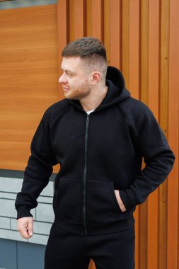 Kendine güvenen genç bir adam, şık bir siyah kapüşonlu giyer, açık havada durur, çarpıcı bir ahşap zemine karşı çağdaş moda seçimlerini vurgular.